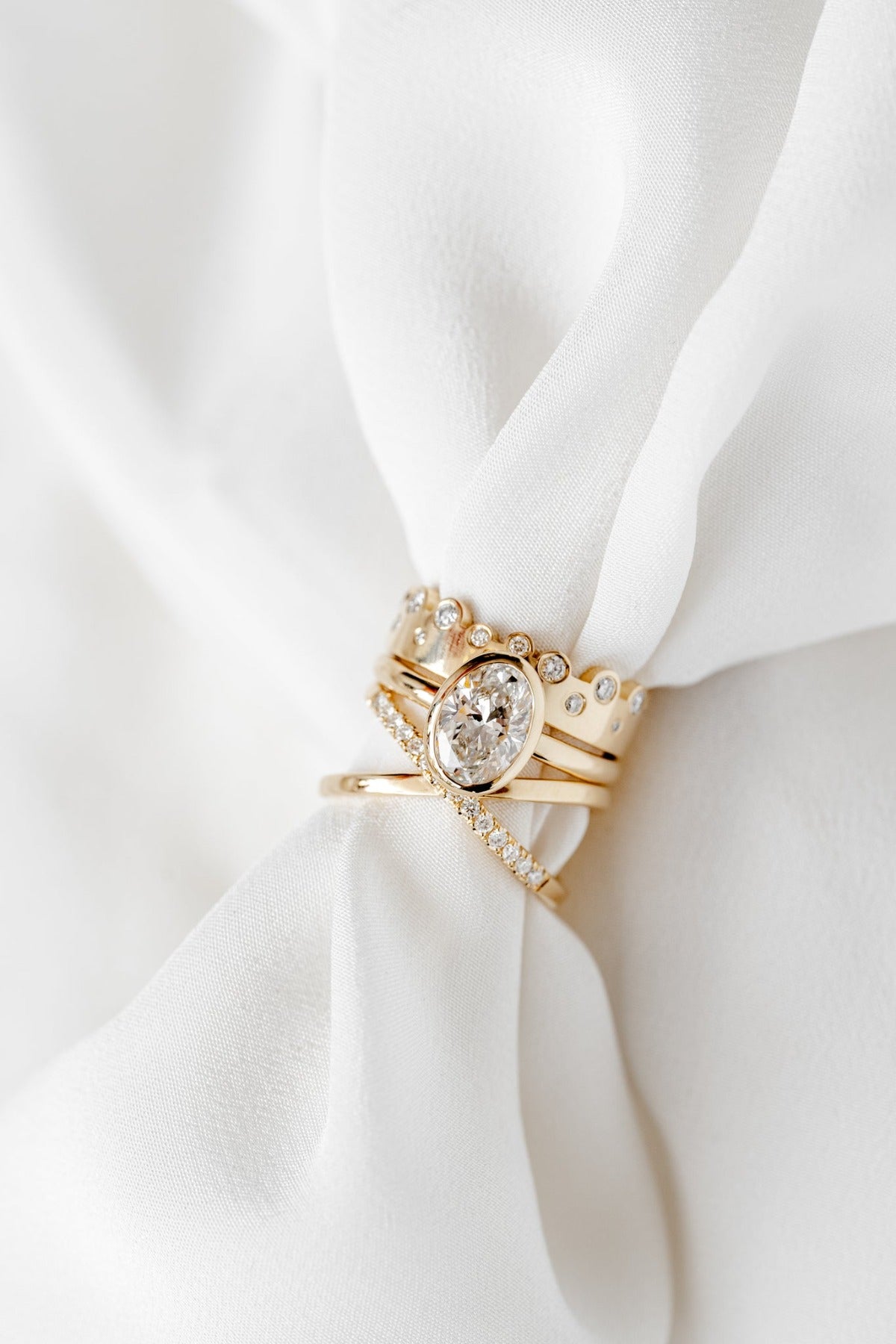 A set of gold rings with diamonds is displayed on a white fabric. The central ring features a large oval-shaped diamond, complemented by smaller diamond-studded bands.