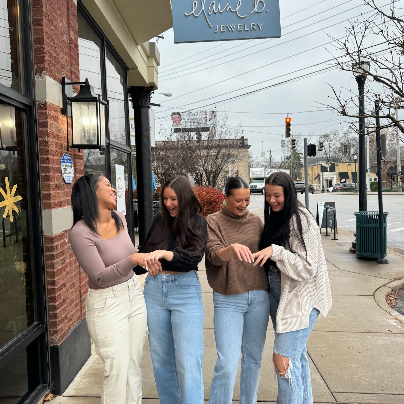 Four women standing in front of Elaine B Jewelry store showing off chain bracelets, smiling and looking at the camera.