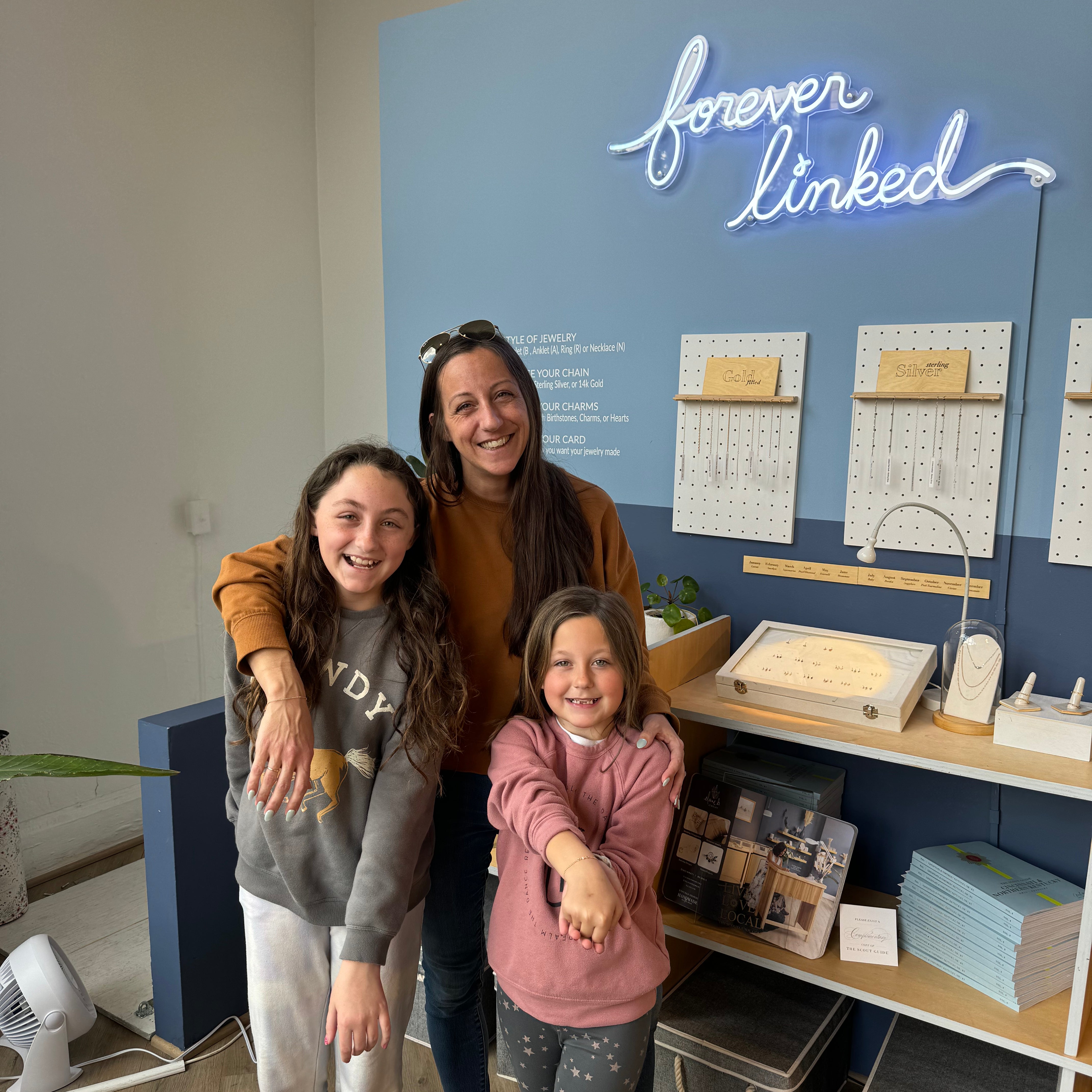 A woman and two girls smiling in front of a sign that reads "forever linked".
