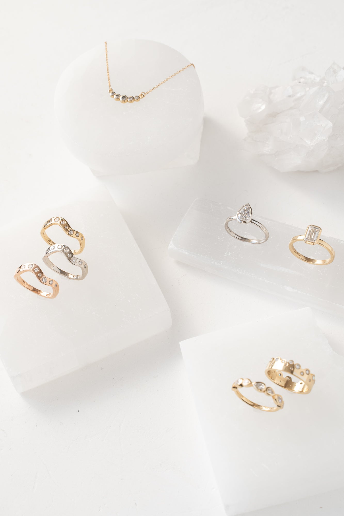Three rings and a necklace on a white surface. The Aura Collection,  features jewelry set in elegant bezels, showcasing a modern and sophisticated design.