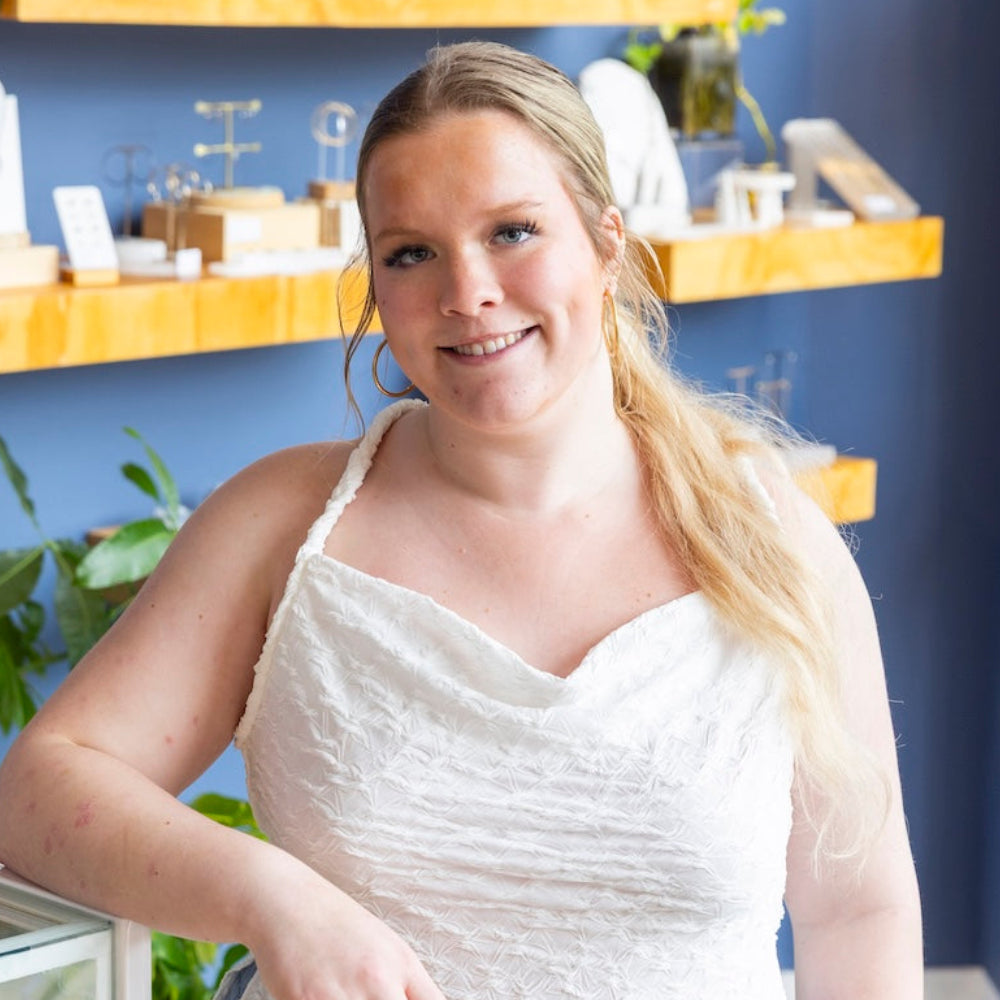 A woman with blonde hair wearing a white textured camisole top and light blue jeans, standing in a jewelry store with blue walls and wooden shelves.