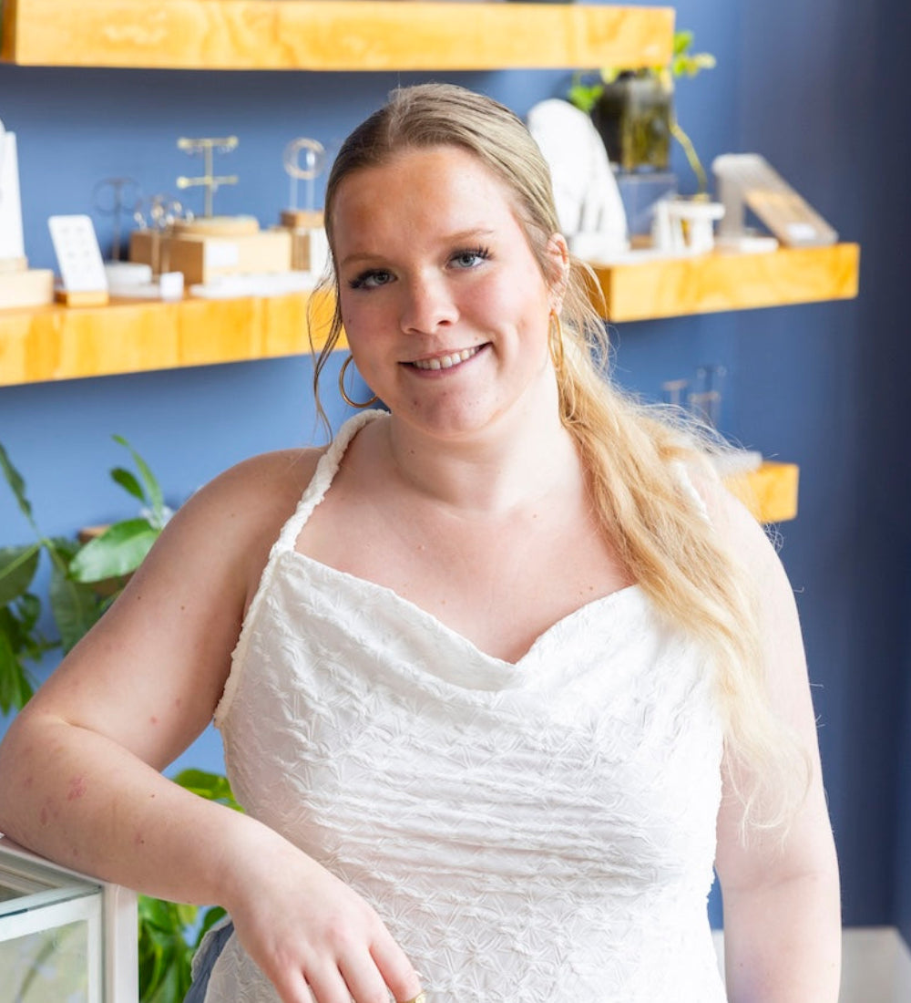 A woman with blonde hair wearing a white textured camisole top and light blue jeans, standing in a jewelry store with blue walls and wooden shelves.