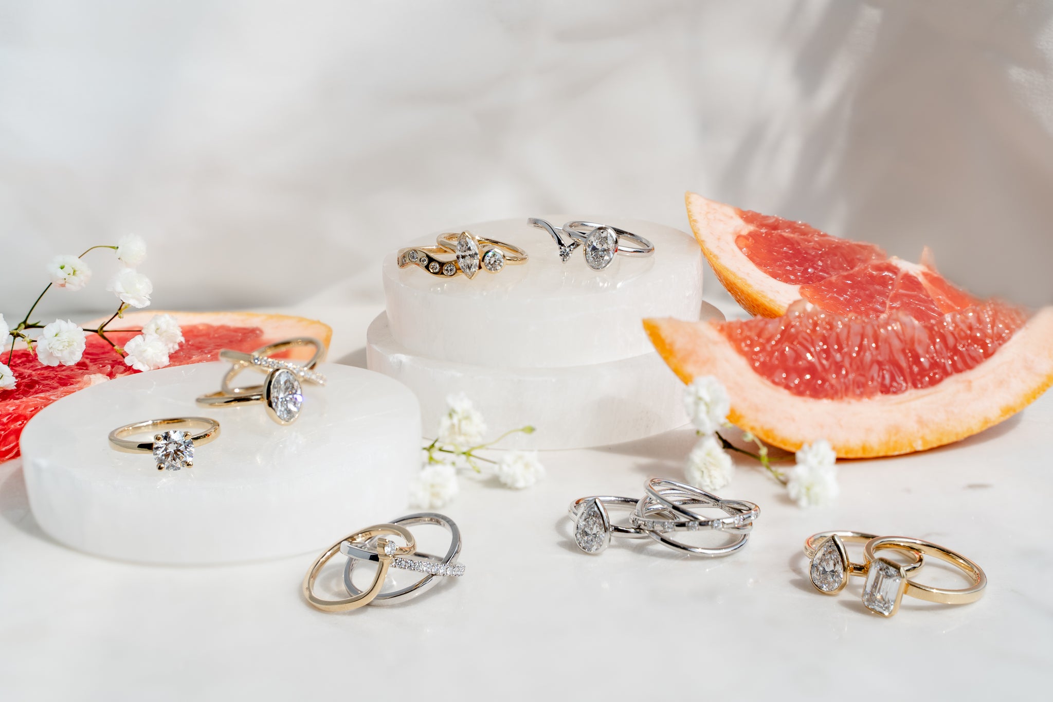 Collection of diamond rings on a white surface with pink grapefruit slices and flowers.