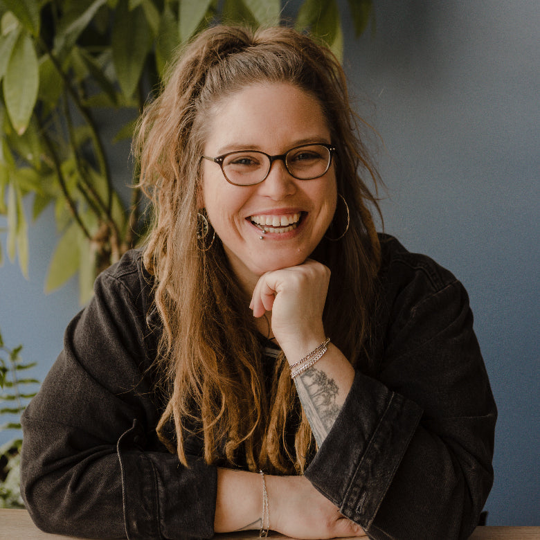 Caitlin, Everyday Production Manager & Senior Jeweler, sitting at a table wearing glasses and a black shirt.