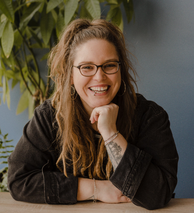 Caitlin, Everyday Production Manager & Senior Jeweler, sitting at a table wearing glasses and a black shirt.
