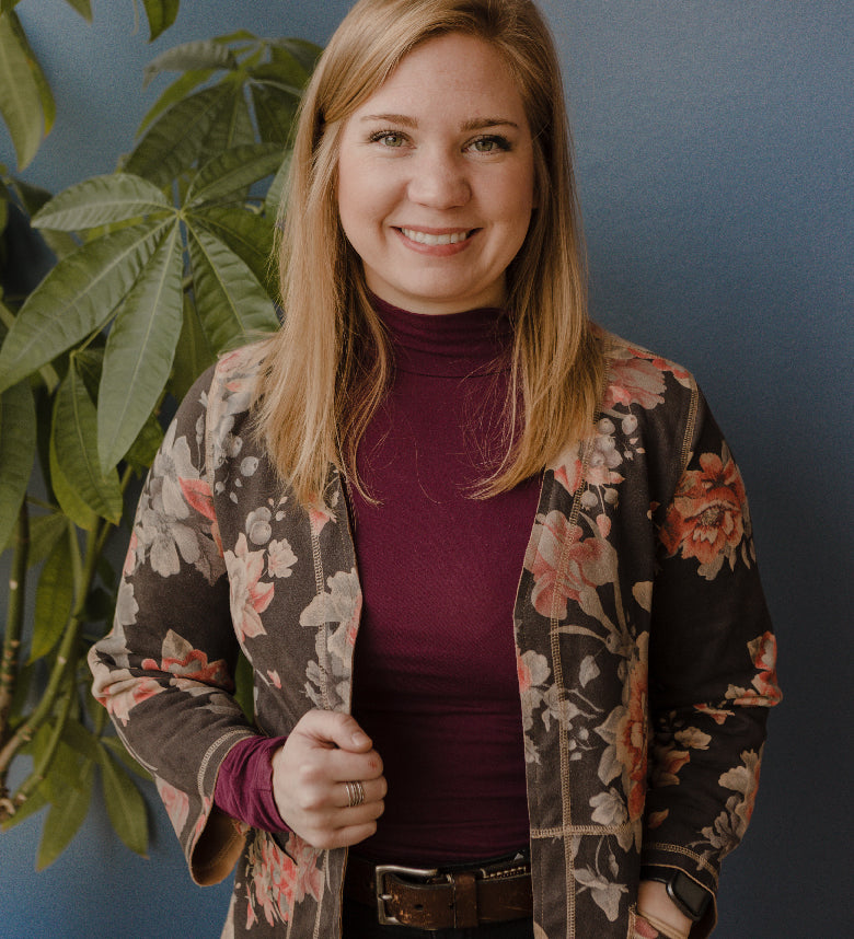 Natasha, a Purchasing Manager & Senior Jeweler, wearing a floral jacket and purple shirt in front a plant and blue wall